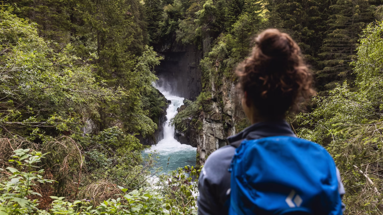 Frau steht im Wald und schaut Richtung eines kleinen Wasserfalls
